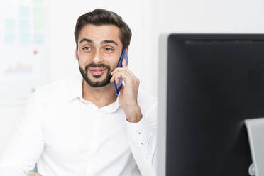 Junger Geschäftsmann, der im Büro sitzend mit seinem Smartphone telefoniert - GIOF10847