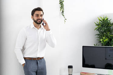 Männlicher Berufstätiger mit Händen in den Taschen bei einem Telefongespräch an einer weißen Wand am Arbeitsplatz - GIOF10848