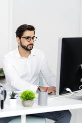 Junger Geschäftsmann mit Brille arbeitet am Computer im Büro - GIOF10853
