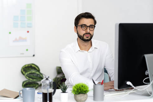 Junger Geschäftsmann mit Brille sitzt vor einem Computer auf einem Schreibtisch am Arbeitsplatz - GIOF10855