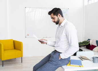 Young businessman with hands in pockets examining paper document in office - GIOF10864