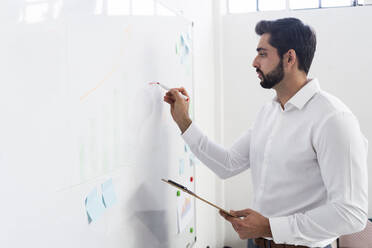 Männlicher Unternehmer, der ein Geschäftsdiagramm auf einem Whiteboard am Arbeitsplatz zeichnet - GIOF10876