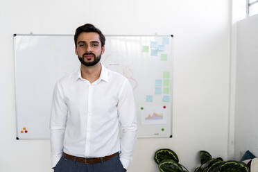 Smiling young businessman with hands in pockets standing against whiteboard at work place - GIOF10883