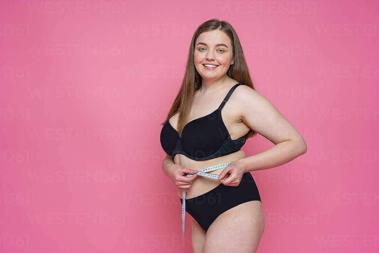 Smiling curvy woman in lingerie measuring abdomen using tape