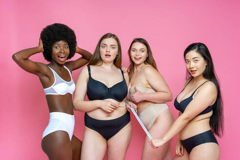 Surprised group of multi-ethnic female model in lingerie with tape measure against pink background - OIPF00138