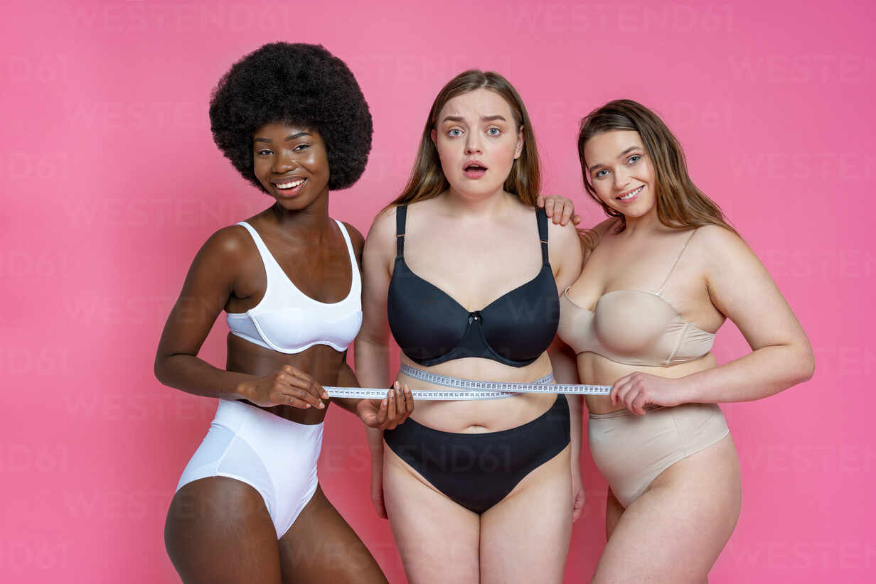 Smiling multi-ethnic female models in lingerie measuring waist of
