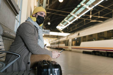 Mann mit Gesichtsschutz, der auf einer Bank im Bahnhof sitzt und ein Mobiltelefon benutzt - EGAF01534