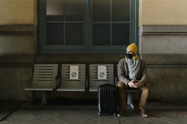 Mann mit Strickmütze und Schutzmaske sitzt mit Gepäck auf einer Bank am Bahnhof - EGAF01535
