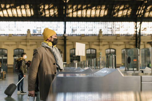 Mann mit Gesichtsschutzmaske geht mit Gepäck durch ein Drehkreuz auf einem Bahnhof - EGAF01536