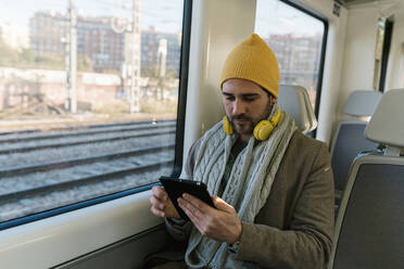 Mittlerer erwachsener Mann in warmer Kleidung, der im Zug sitzend ein digitales Tablet benutzt - EGAF01537