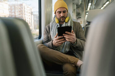 Mann mit Strickmütze und Kopfhörer, der im Zug sitzend ein digitales Tablet benutzt - EGAF01538