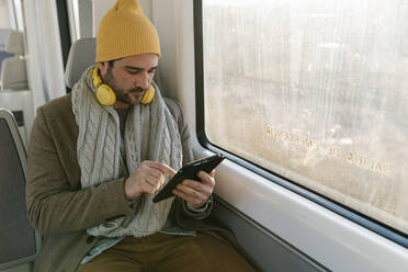 Mittlerer erwachsener Mann mit Strickmütze und Kopfhörern, der im Zug sitzend ein digitales Tablet benutzt - EGAF01539