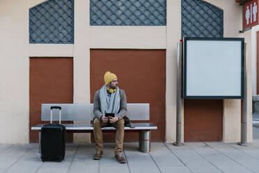 Mittlerer Erwachsener mit Strickmütze, der ein digitales Tablet benutzt, während er auf einer Bank am Bahnhof sitzt - EGAF01542