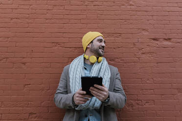 Modischer Mann, der ein digitales Tablet benutzt, während er sich an eine Backsteinmauer lehnt - EGAF01543