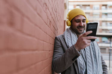Man with headphones smiling while using mobile phone leaning on wall - EGAF01545