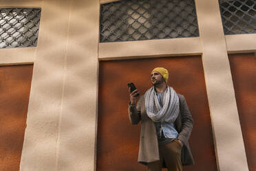 Modischer Mann mit Händen in den Taschen, der ein Mobiltelefon benutzt, während er an der Wand steht - EGAF01547