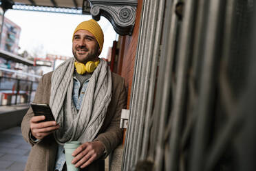 Lächelnder Mann mit Kaffeetasse und Mobiltelefon, der wegschaut, während er auf dem Bahnhof steht - EGAF01556