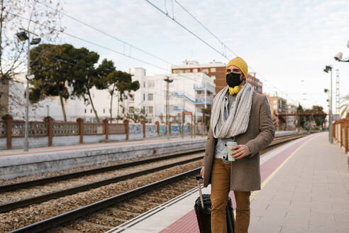 Mann mit Schutzmaske hält Kaffeetasse und Gepäck beim Gehen auf dem Bahnhof - EGAF01558