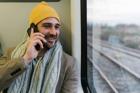 Lächelnder Mann mit Strickmütze, der im Zug sitzend mit seinem Mobiltelefon spricht - EGAF01562