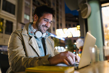 Lächelnder Mann, der einen Laptop benutzt, während er in einem Straßencafé sitzt - EGAF01572