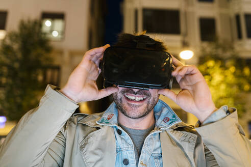 Mittelgroßer erwachsener Mann, der ein Virtual-Reality-Headset trägt, während er im Freien sitzt - EGAF01578