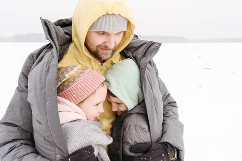 Vater, der seine Kinder in seinen Mantel hüllt, während er im Winter an Land steht - EYAF01497