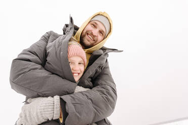 Cheerful father covering daughter in winter coat against clear sky - EYAF01498