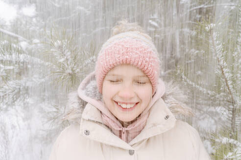 Nahaufnahme eines lächelnden Mädchens mit geschlossenen Augen, das den Schneefall im Wald genießt - EYAF01509