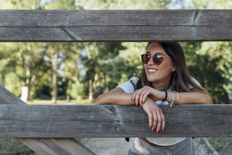 Smiling young woman wearing sunglasses leaning on wooden fence in forest stock photo