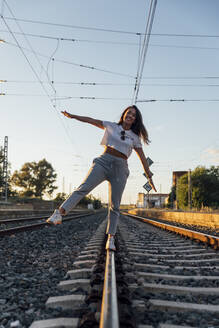Lächelnde junge Frau mit ausgestreckten Armen, die auf einem Bahngleis gegen den klaren Himmel läuft - JRVF00178
