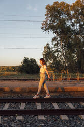 Carefree woman with arms outstretched walking on railroad track against clear sky - JRVF00179