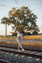 Carefree young woman with arms outstretched walking on railroad track during sunset - JRVF00180