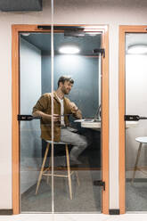 Male professional talking on video call through laptop seen through glass door in office - VPIF03417