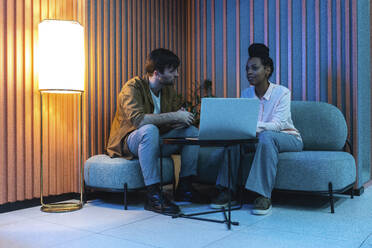 Männliche und weibliche Kollegen mit Laptop, die an einem beleuchteten Arbeitsplatz sitzen und planen - VPIF03430