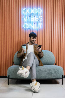 Smiling creative businessman with coffee cup in unicorn slippers using smart phone against wall in office - VPIF03432