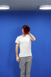 Kreativer Geschäftsmann schaut durch ein Megaphon, während er an einer blauen Wand am Arbeitsplatz steht - VPIF03458
