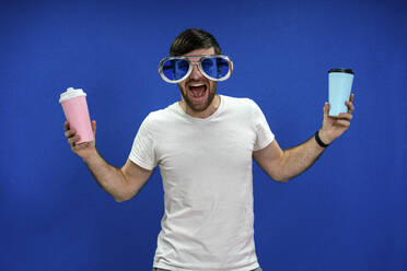 Cheerful businessman with mouth open in oversized sunglasses holding reusable coffee cups against blue wall in office - VPIF03462