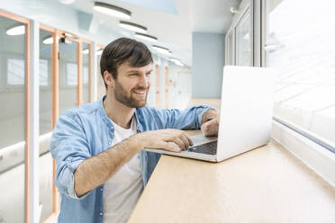 Smiling businessman using laptop on window seat at office - VPIF03482