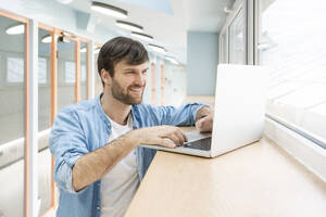 Lächelnder Geschäftsmann mit Laptop auf dem Fensterplatz im Büro - VPIF03482