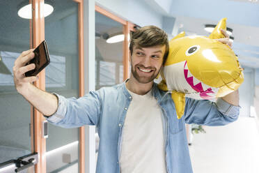 Lächelnder kreativer Geschäftsmann, der ein Selfie mit einem Ballonhai im Korridor am Arbeitsplatz macht - VPIF03488