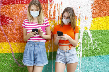 Fashionable female friends in shorts using smart phones against graffiti wall during pandemic - IFRF00340