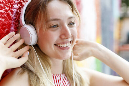 Lächelnde junge Frau, die wegschaut, während sie mit Kopfhörern an einer bunten Wand Musik hört - IFRF00345