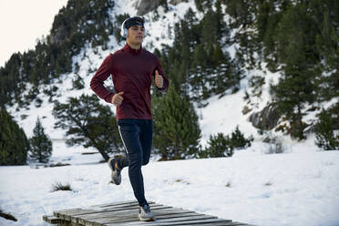 Entschlossener junger männlicher Sportler, der im Winter auf einem Steg läuft und dabei Musik hört - ACPF01059