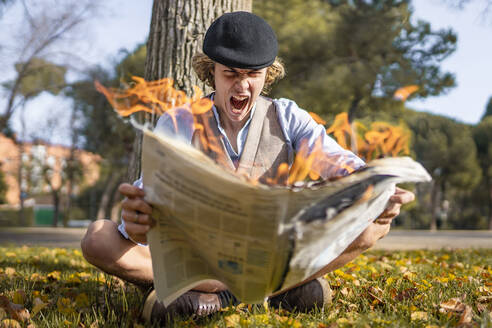 Screaming young man looking at lit newspaper while sitting in park - GGGF00923