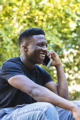 Lächelnder junger Mann, der im Park sitzt und mit seinem Smartphone spricht - PNAF00579