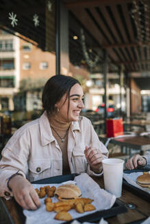 Fröhliches Teenager-Mädchen, das seinen Freund anschaut, während es in einem Restaurant im Freien Fastfood isst - GRCF00630