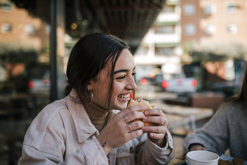 Close-up of cheerful teenage girl eating burger with friend at sidewalk cafe - GRCF00634