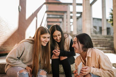 Cheerful teenage girl showing smart phone to friends while sitting on steps in city - GRCF00635