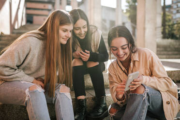 Smiling teenage girl showing mobile phone to friends while sitting on steps in city - GRCF00636