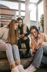Smiling teenage girl showing mobile phone to friends while sitting on steps - GRCF00637